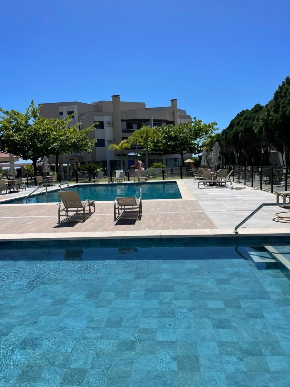 Piscina adulta e infantil do condomínio em dia de sol, com espreguiçadeiras e o prédio ao fundo